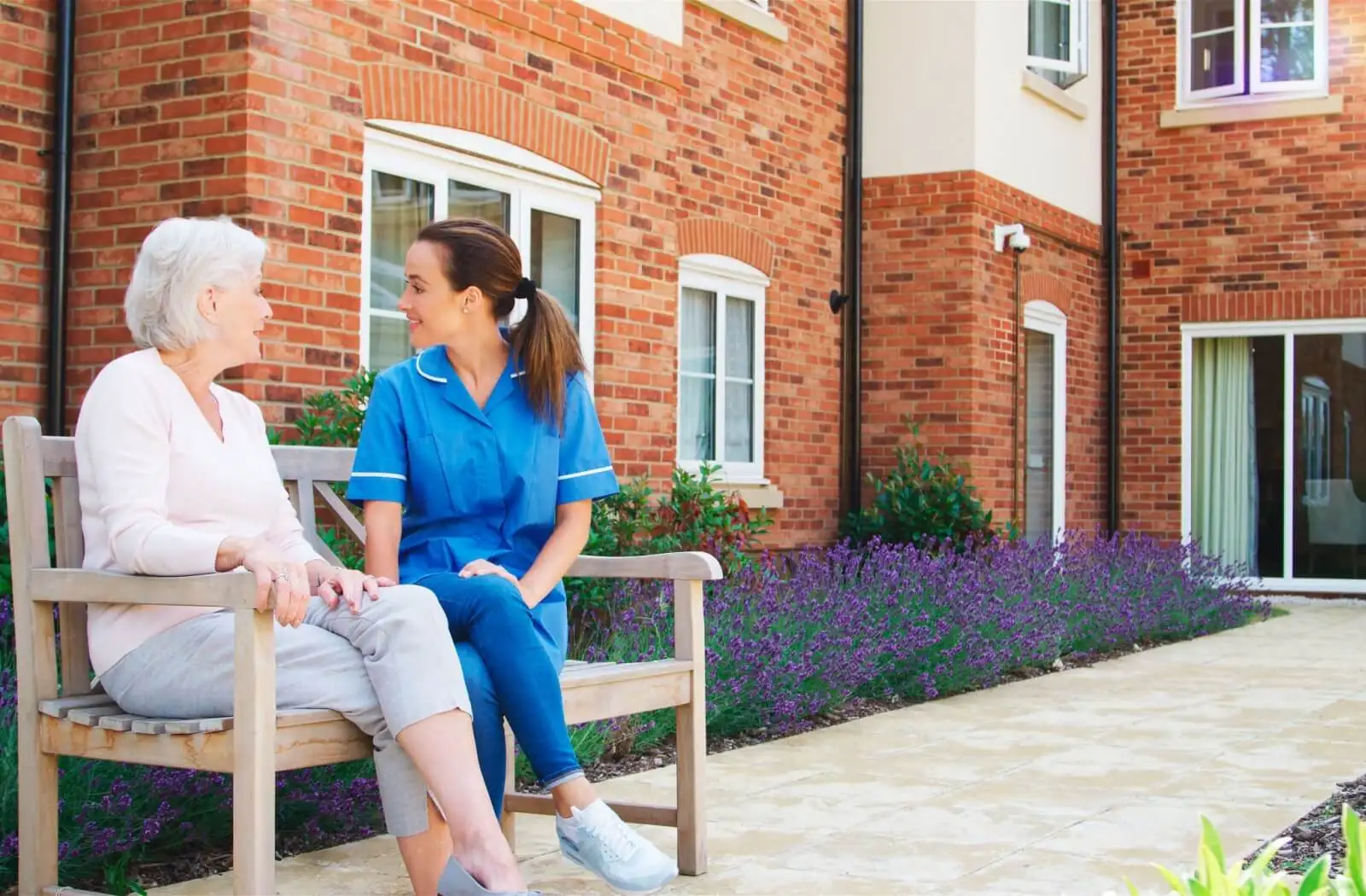 Caregiver and elderly woman enjoying a conversation on a bench in a garden setting, featuring a brick building and blooming lavender. Ideal for highlighting senior care and companionship.