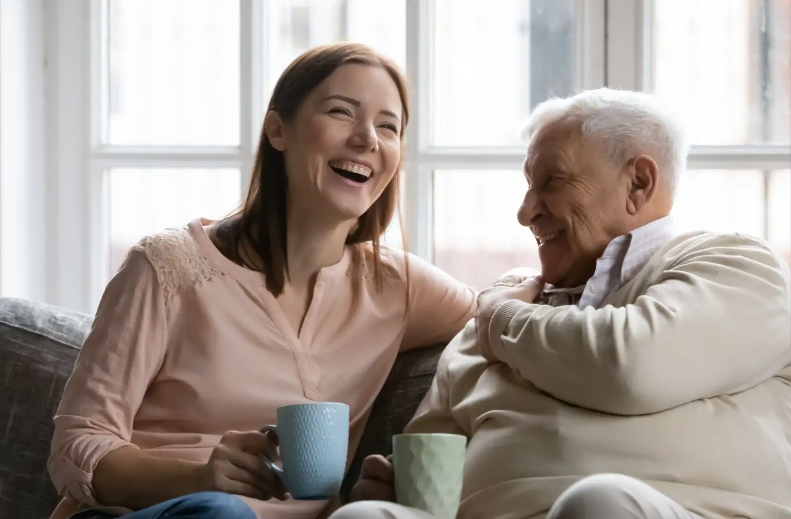 A woman and an elderly man share a joyful moment while drinking tea in a cozy living room, highlighting the warmth of intergenerational relationships.