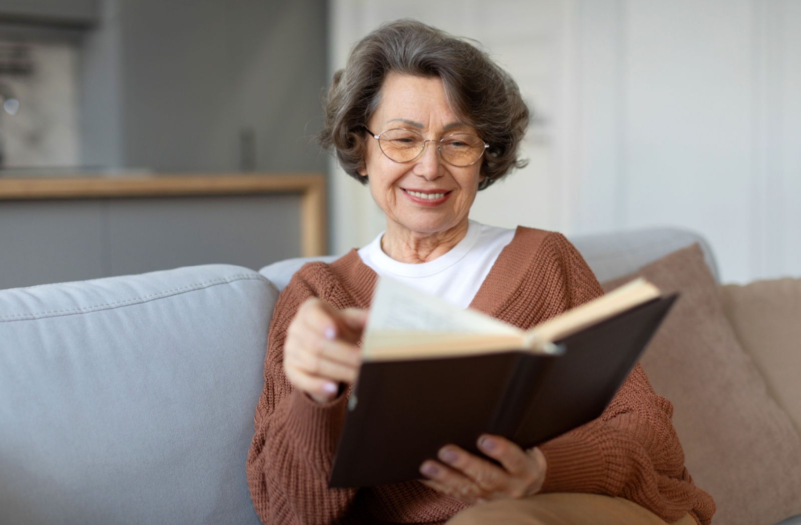 A senior woman reads a book on a couch.