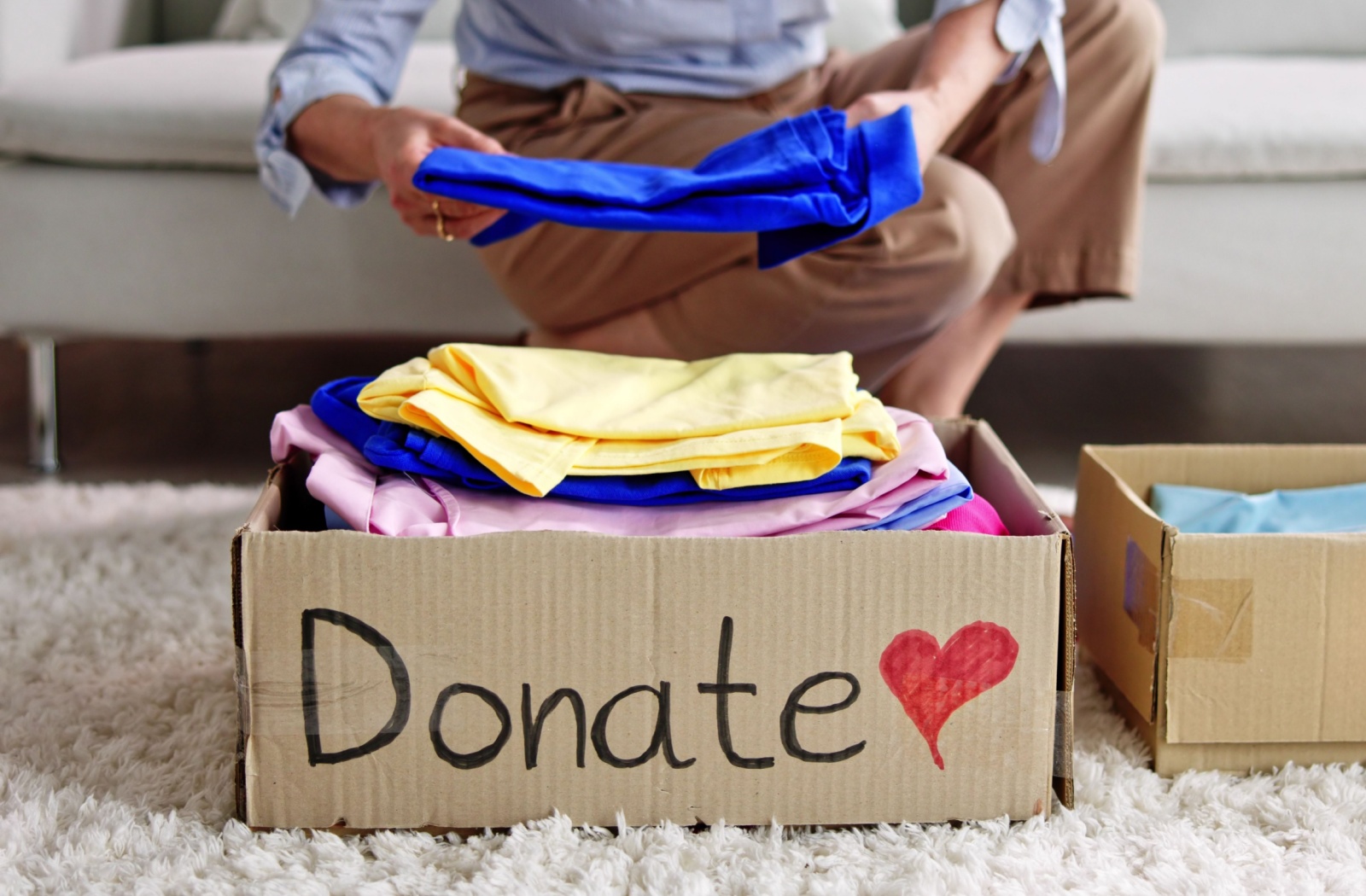 An older adult places several brightly colored pieces of cloth into a box labelled “Donate” as a part of downsizing